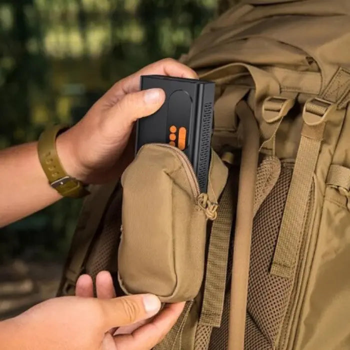 Hands placing small black and orange device into a tan backpack pocket, demonstrating portability