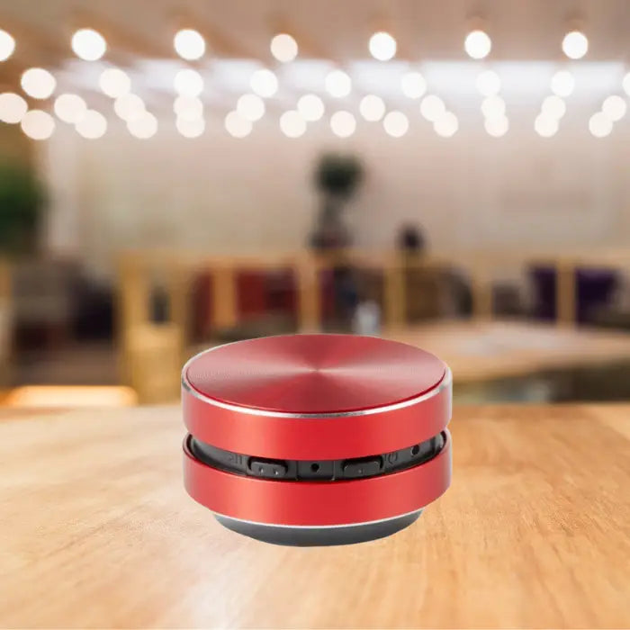 compact red bluetooth speaker with metallic finish displayed on wooden table with ambient bokeh lighting effect