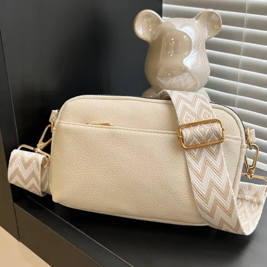 Cream crossbody bag with trendy chevron strap and gold accents, perfect for modern casual outfits.