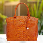 Crocodile embossed handbag in bright orange with textured body and short handles on white table background.