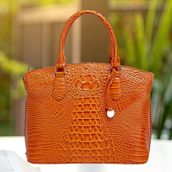 Crocodile embossed handbag in bright orange with textured body and short handles on white table background.