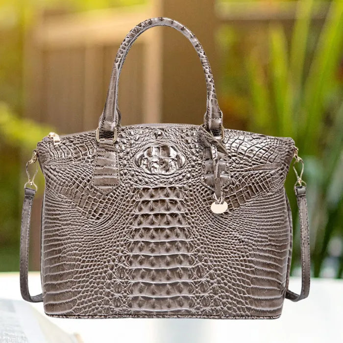 Crocodile embossed handbag in dark taupe with gold hardware and zip top, shown outdoors with blurred greens.