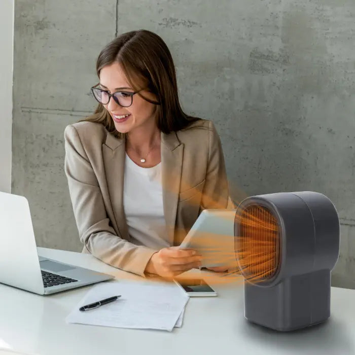 Desktop electric heater in use by businesswoman at office desk for personal warmth.