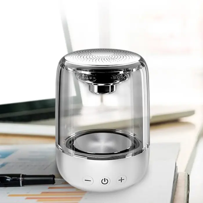 Transparent wireless speaker with silver top on white base, placed on desk with laptop, papers, and pen