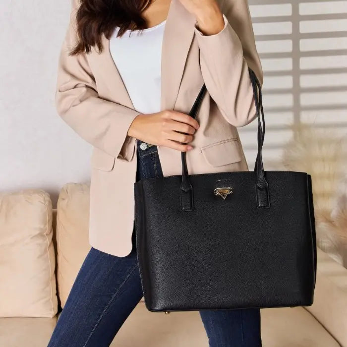 Woman wearing beige blazer and jeans holding a structured black leather tote bag with gold hardware against a light background