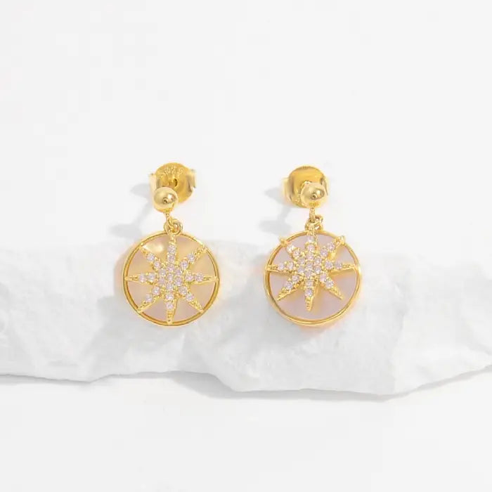 Gold starburst drop earrings with crystal details displayed on white textured surface with soft shadows