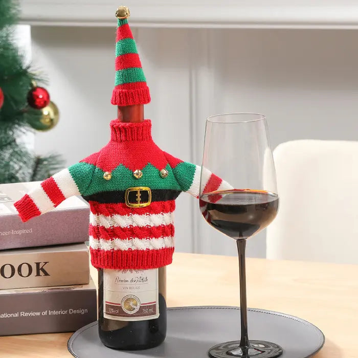 Striped elf-themed wine bottle cover with bells and pointed hat displayed with books and wine glass