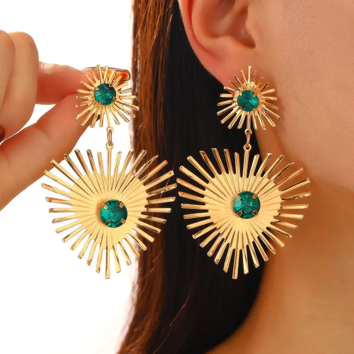 emerald sunburst statement earrings in gold with large green gems shown close to ear, featuring bold radiant lines for standout style