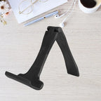 Foldable phone stand in black shown open on wooden desk with accessories around it for workspace use.