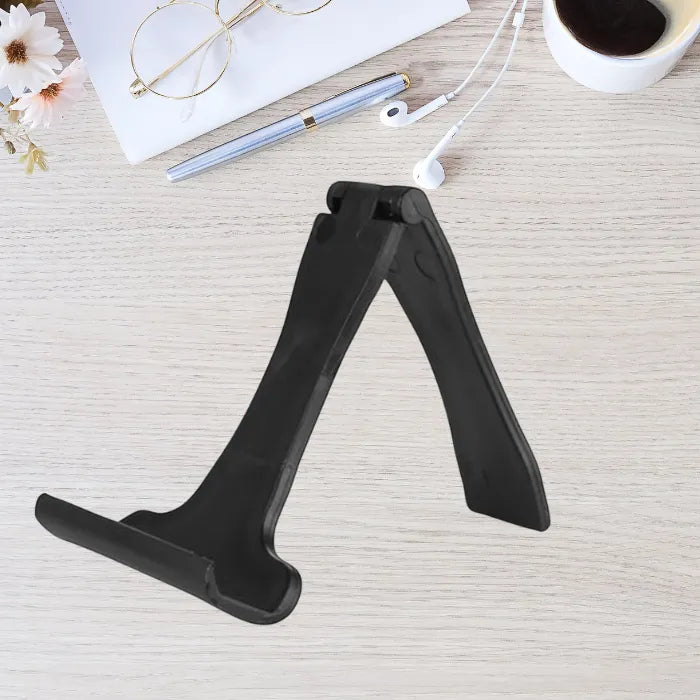 Foldable phone stand in black shown open on wooden desk with accessories around it for workspace use.