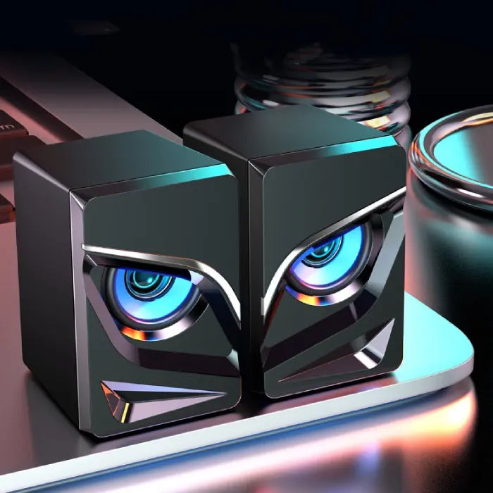 Futuristic black computer speakers with neon blue eye-like lights on reflective surface with colorful lighting