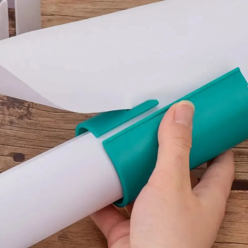 Gift wrap cutter in teal being used by hand to slice white wrapping paper over wooden surface.