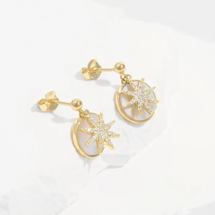 Gold plated starburst earrings with crystal pavé details and circular mother of pearl backdrop on white surface