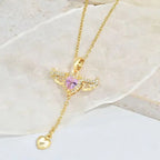 Gold heart pendant necklace with pink gemstone and wing design, placed on a soft white backdrop.