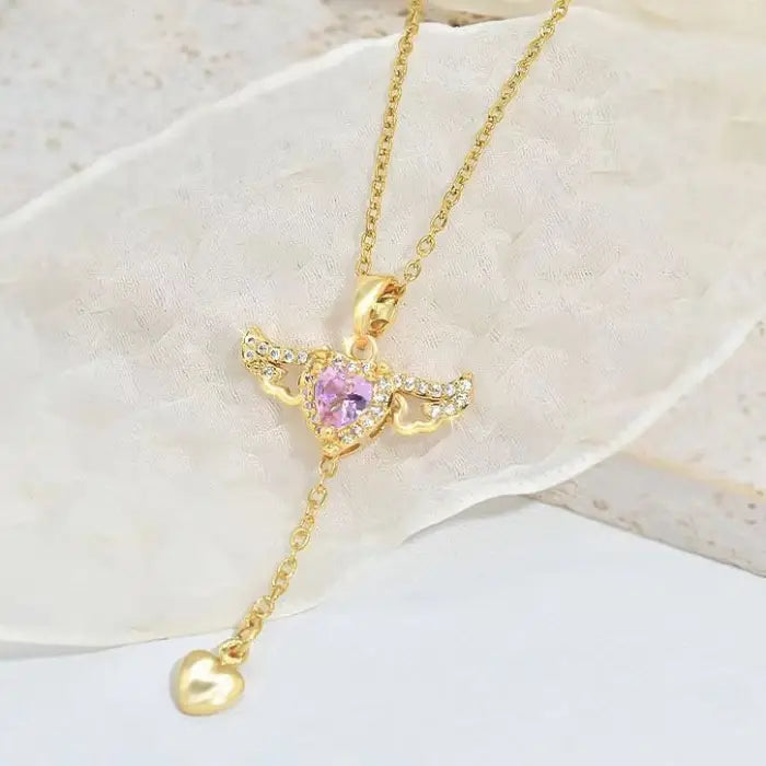 Gold heart pendant necklace with pink gemstone and wing design, placed on a soft white backdrop.