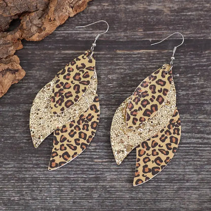 Layered leaf-shaped earrings featuring leopard print and glitter accents hanging from a rustic stick with a blurred background.