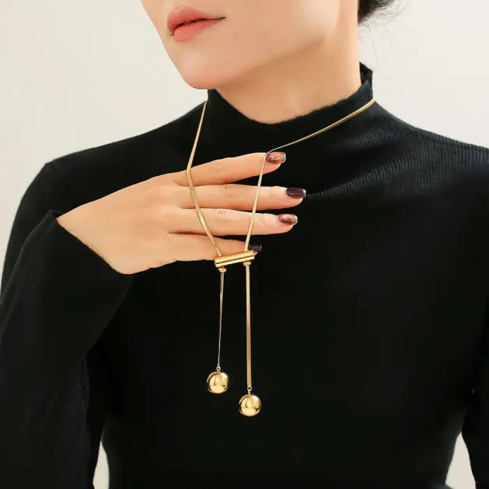 Hand holding gold pendant necklace against black sweater, showcasing dual hanging spheres and adjustable design