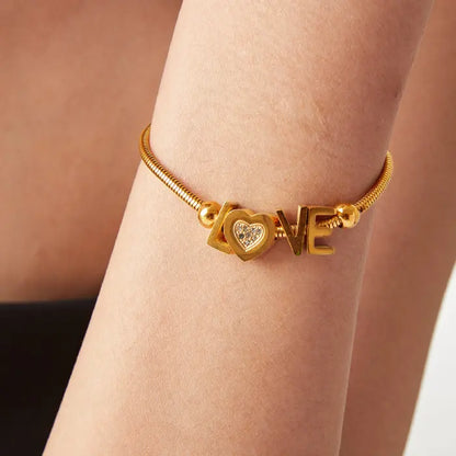 Close-up of gold LOVE bracelet worn on wrist, showing crystal heart detail and snake chain design