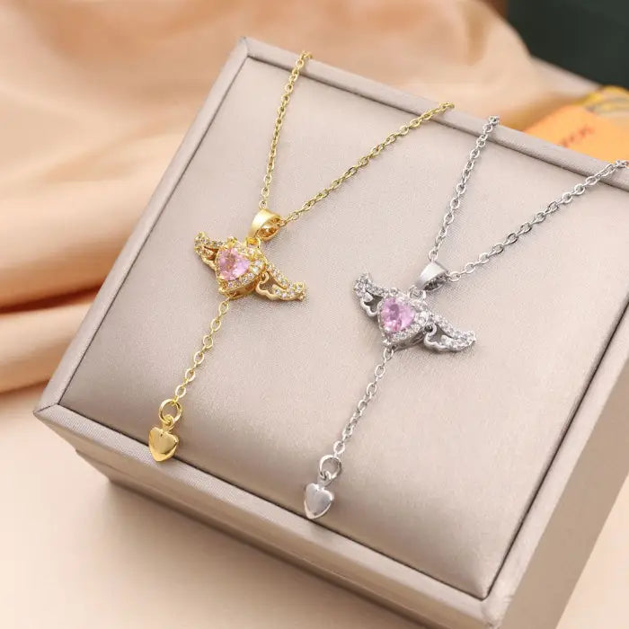 Gold and silver heart pendant necklaces with wing designs and pink gemstone, displayed on a box.