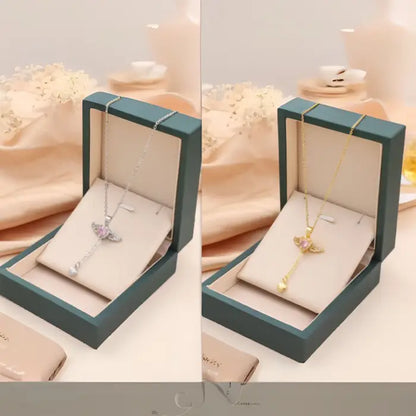 Gold and silver heart necklaces with wing designs and pink gemstones, displayed side by side in boxes.