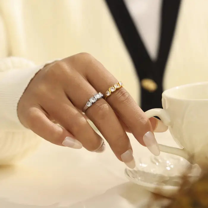 Hand wearing gold and silver cubic zirconia rings holding a white coffee cup, styled in a cozy setting.