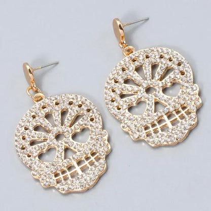 Close-up of gold skull-shaped dangle earrings with clear rhinestones on a gray background.