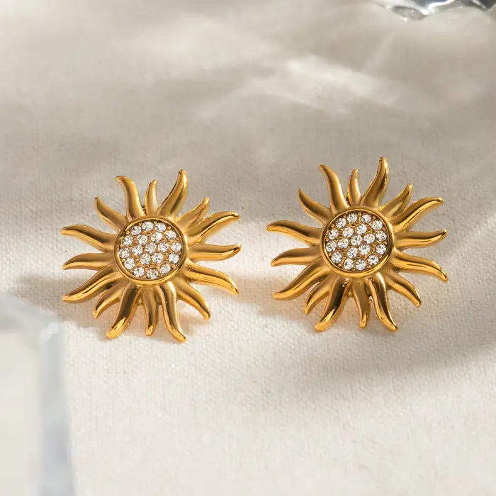 Gold sun design earrings with diamond-like details on a textured fabric background.