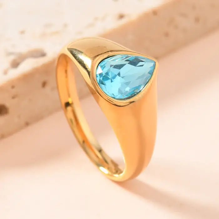 gold teardrop birthstone ring with brilliant blue gemstone in smooth gold band, photographed against soft tones to highlight sparkle