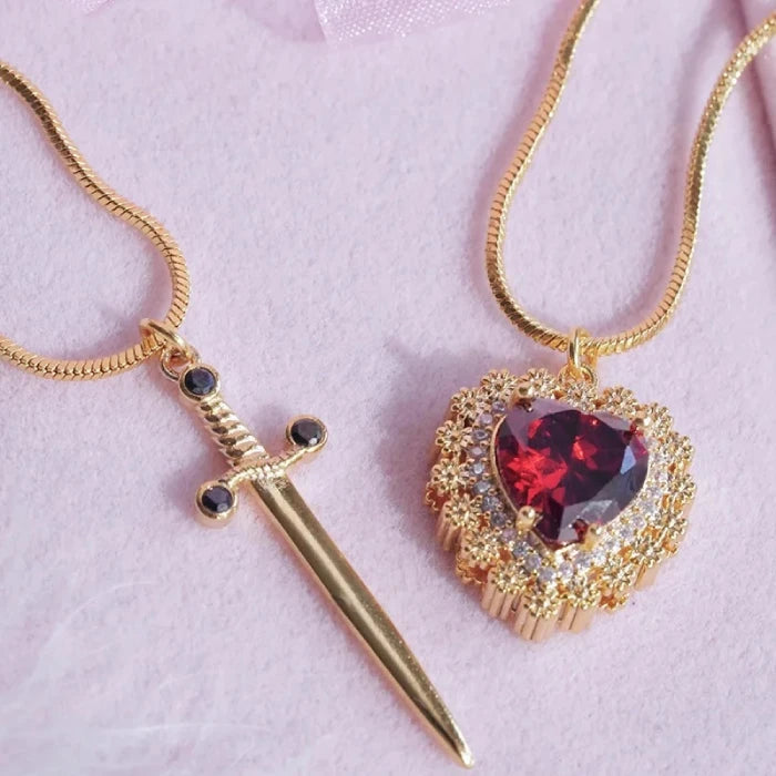 Gothic Heart Necklace in gold with floral frame around deep red gemstone on pink background.