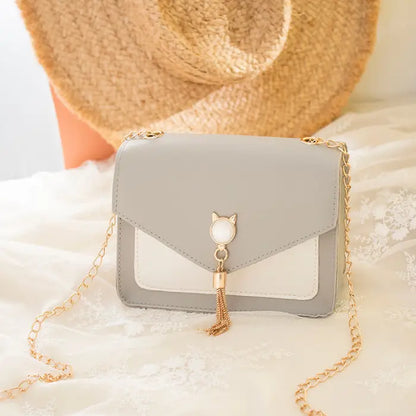 Gray crossbody bag with gold chain, cat charm, and tassel detail on lace fabric with a straw hat background.