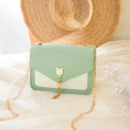 Green crossbody bag with gold chain, cat charm, and tassel detail on lace fabric with a straw hat backdrop.