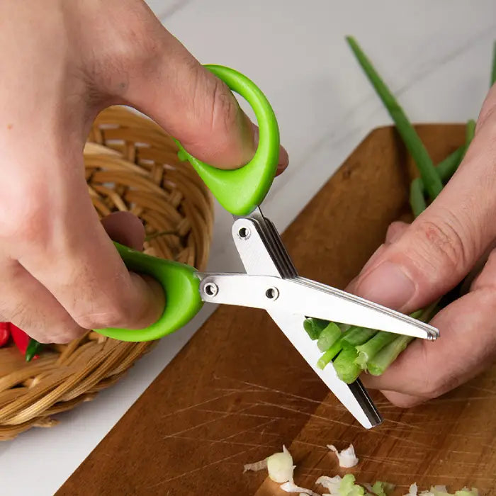 Multi-blade herb scissors with green handle cutting fresh green onions on a wooden cutting board.