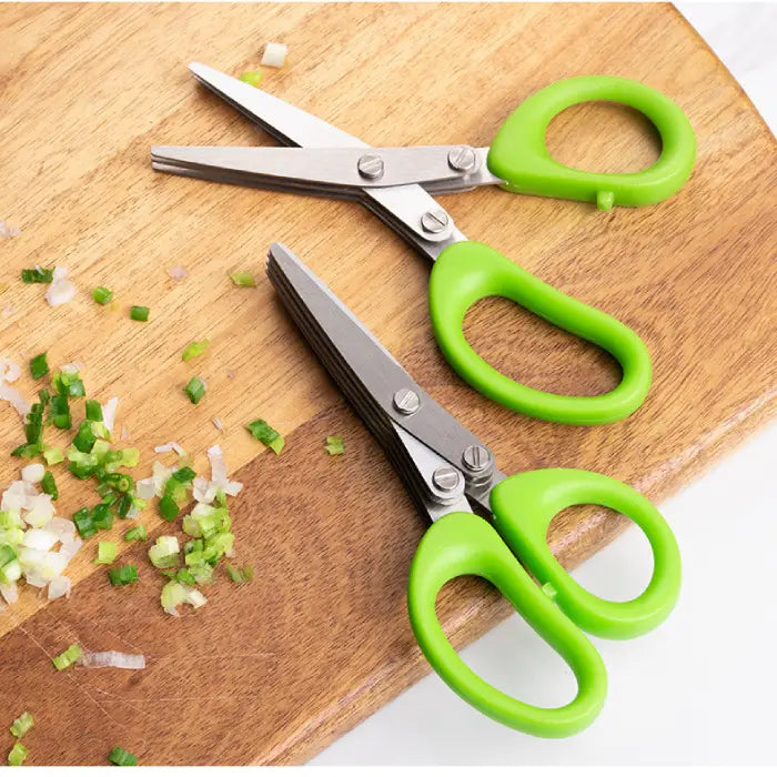 Two green-handled multi-blade scissors on a wooden board surrounded by chopped green onions.