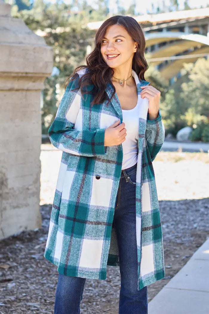 Woman showing the front view of a green plaid wool coat, styled casually with jeans in an outdoor park setting.