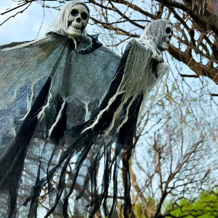 Floating halloween skeleton ghosts with tattered black cloth hanging from tree branches in daylight.
