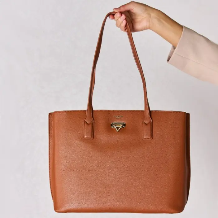 Hand holding brown leather tote bag with gold hardware against white background, showcasing bag's structure and details