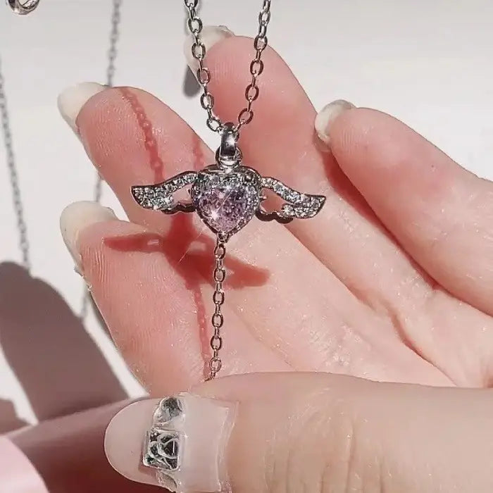 Close-up of a hand holding a silver heart pendant necklace with wing design and pink gemstone.