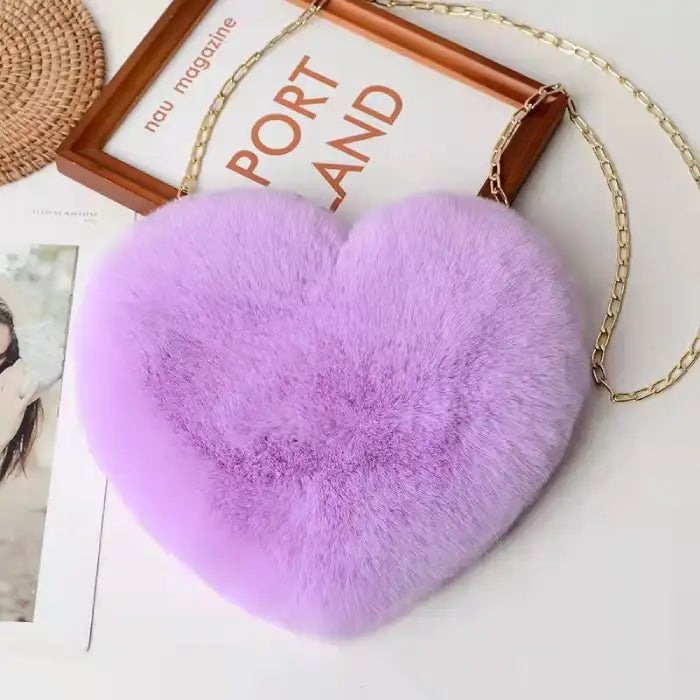 Heart shoulder bag in lavender fluffy plush with gold chain, placed on a magazine-themed background.