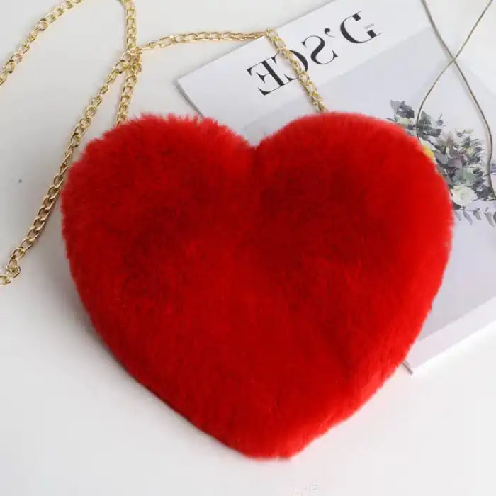 Red heart-shaped shoulder bag in plush finish, flat on surface with romantic magazine accent.