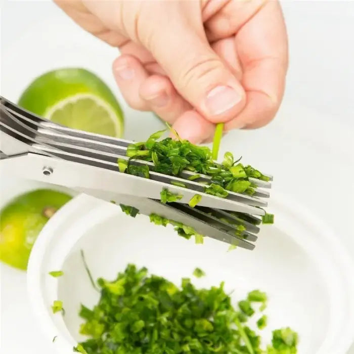 Herb scissors cutting fresh herbs into a bowl, with lime in the background on a white surface.