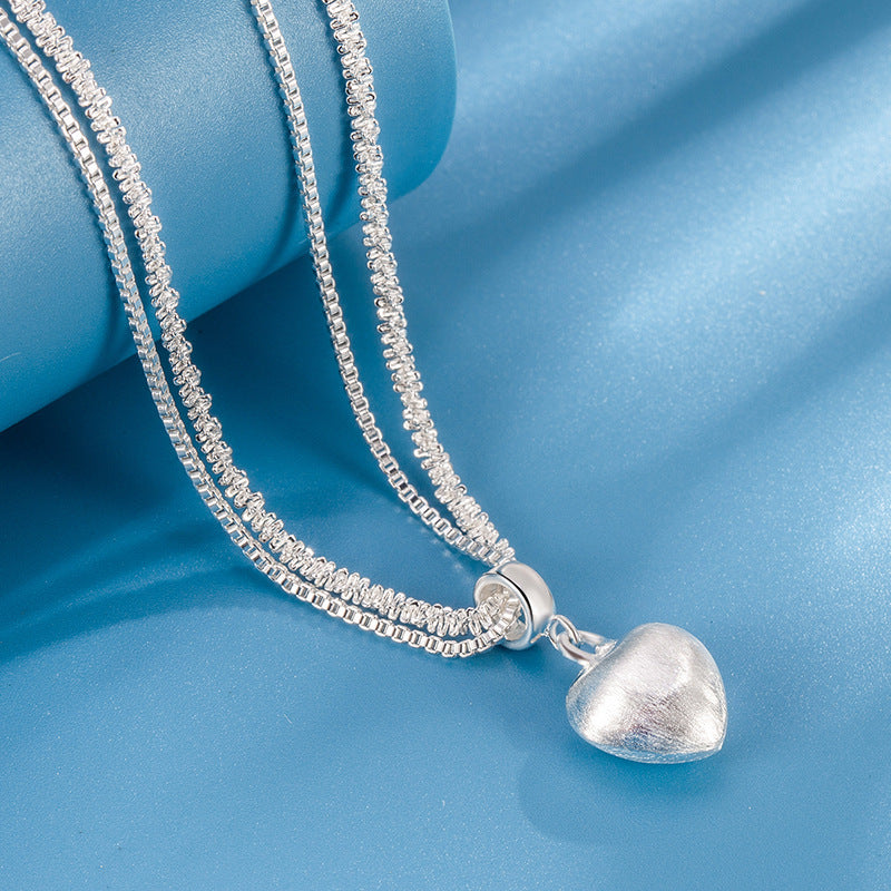 layered silver heart necklace with brushed heart pendant draped on blue surface showing chain texture clearly