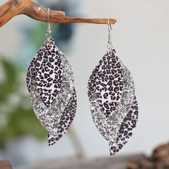 Stylish layered earrings with leopard print and glitter details hanging from a wooden branch with a blurred background.