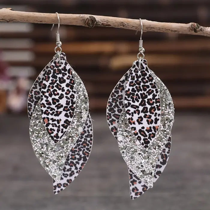 Layered leaf-shaped earrings featuring leopard print and glitter accents hanging from a rustic stick with a blurred background.