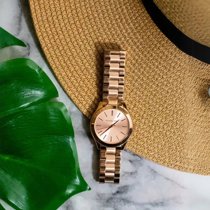 Rose gold Michael Kors watch displayed on marble surface with straw hat and monstera leaf in lifestyle setting