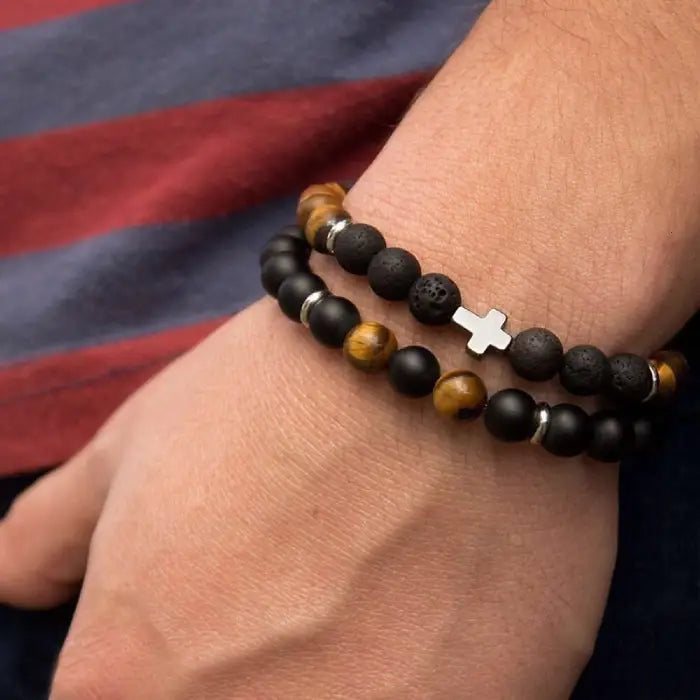 mens bracelet stack. Close-up of a wrist wearing a beaded bracelet with a cross charm against a striped fabric background.