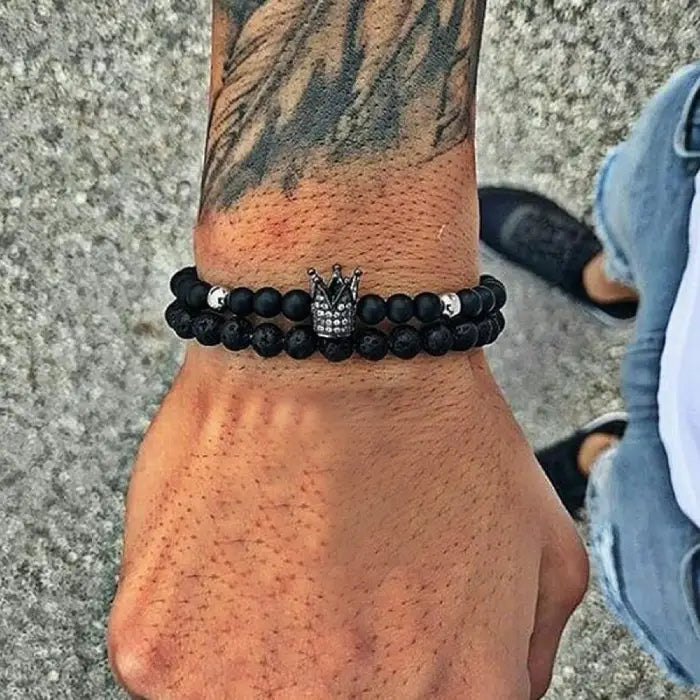 mens bracelet stack. Black beaded bracelet with a crown charm on a wrist with tattoos, against a concrete floor background.