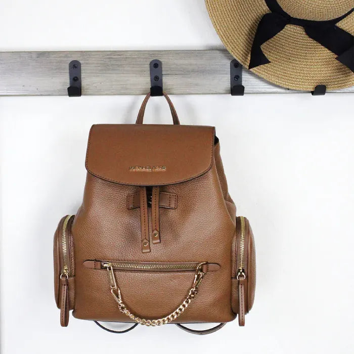 Michael Kors brown leather backpack hanging on hooks, paired with a straw hat for stylish display.