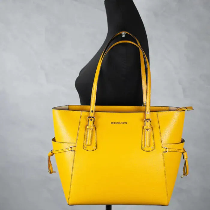 Yellow Michael Kors Voyager tote displayed on black mannequin against grey background.