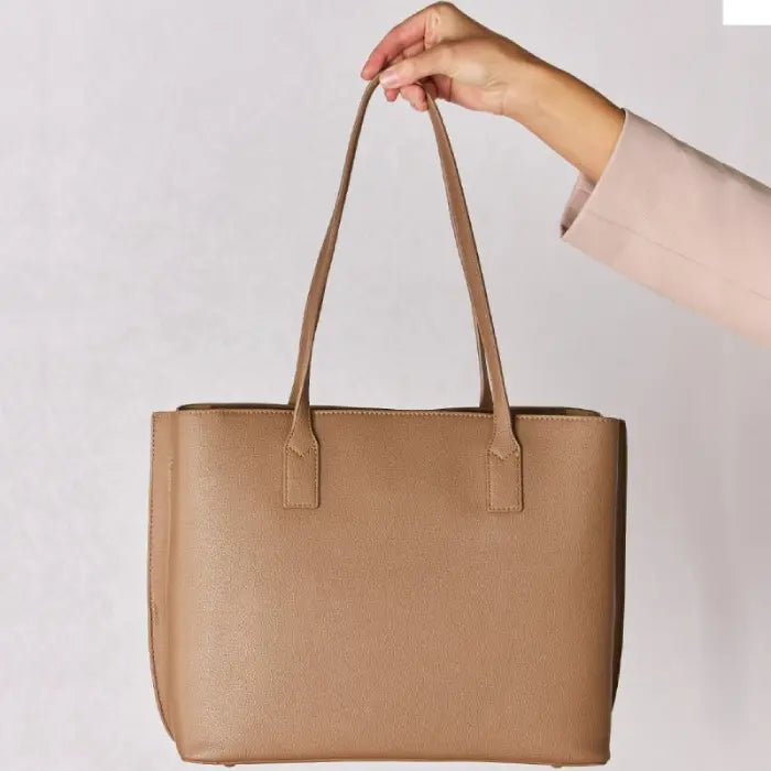 Hand holding a simple, elegant tan leather tote bag against a white background, highlighting its clean lines and shape