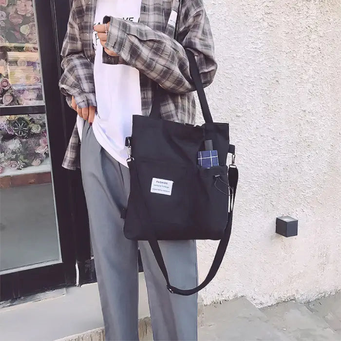 Black canvas tote with adjustable strap shown with casual gray pants and plaid shirt